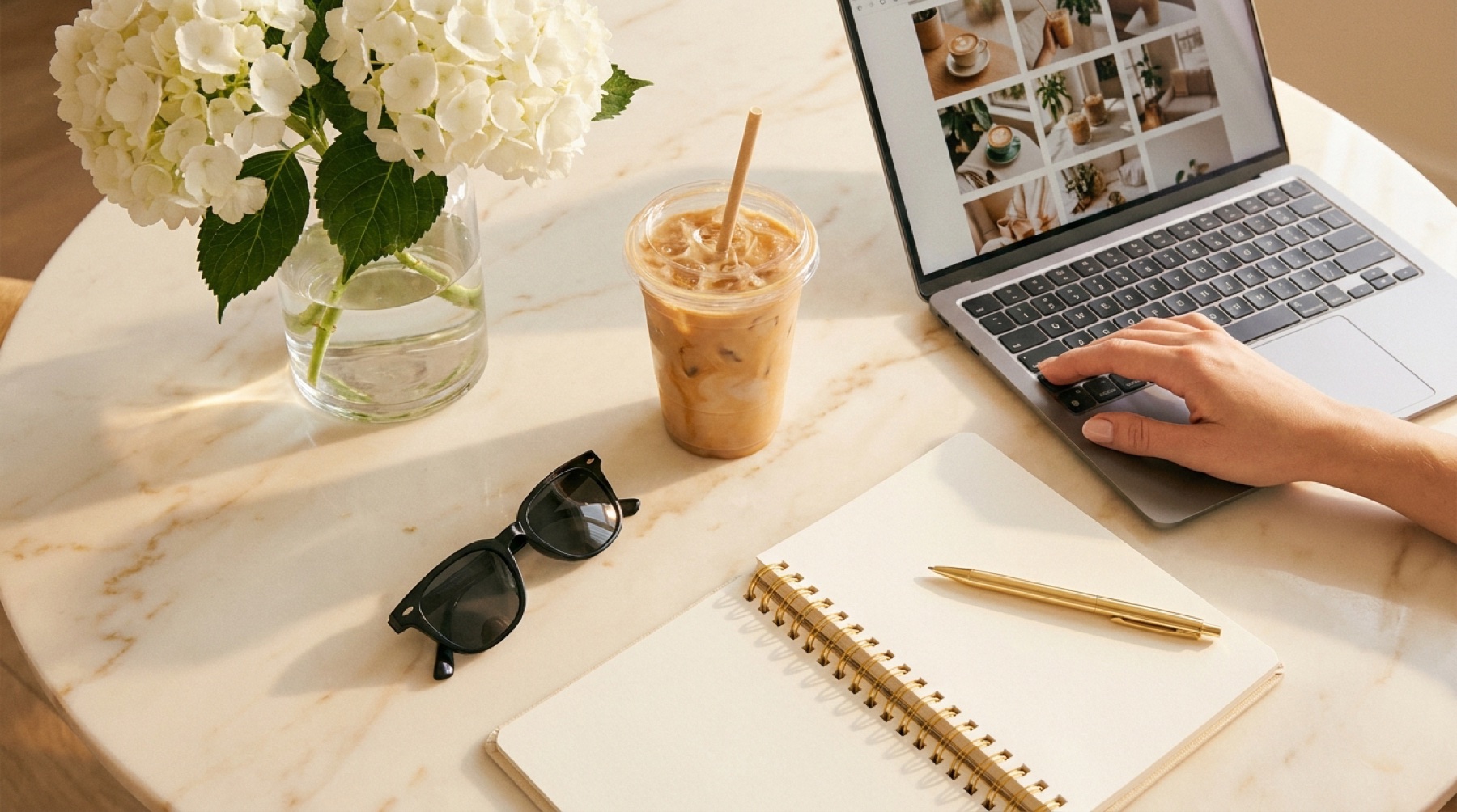 Fresh white hydrangeas, an iced latte, sunglasses and an open gold-spiral notebook on a marble desk beside a laptop